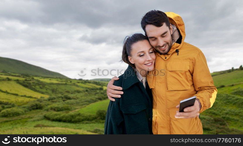 technology, travel and tourism concept - happy couple with smartphone over farmland fields and hills at wild atlantic way in ireland. couple with smartphone in ireland