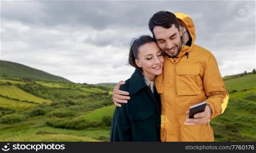 technology, travel and tourism concept - happy couple with smartphone over farmland fields and hills at wild atlantic way in ireland. couple with smartphone in ireland