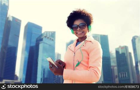 technology, travel, tourism and people concept - smiling african american young woman or teenage girl with smartphone and headphones listening to music over singapore city background