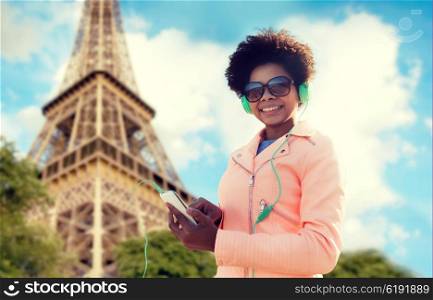 technology, travel, tourism and people concept - smiling african american young woman or teenage girl with smartphone and headphones listening to music over eiffel tower background