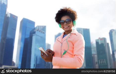 technology, travel, tourism and people concept - smiling african american young woman or teenage girl with smartphone and headphones listening to music over singapore city background