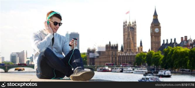 technology, travel, tourism and people concept - smiling young man or teenage boy in headphones with smartphone listening to music over london city and big ben tower background