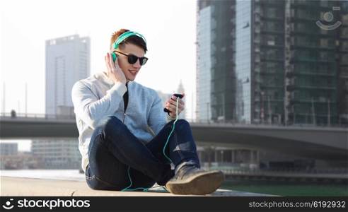 technology, travel, tourism and people concept - smiling young man or teenage boy in headphones with smartphone listening to music over dubai city street background