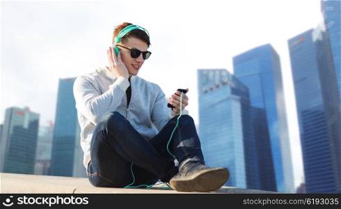 technology, travel, tourism and people concept - smiling young man or teenage boy in headphones with smartphone listening to music over singapore city skyscrapers background