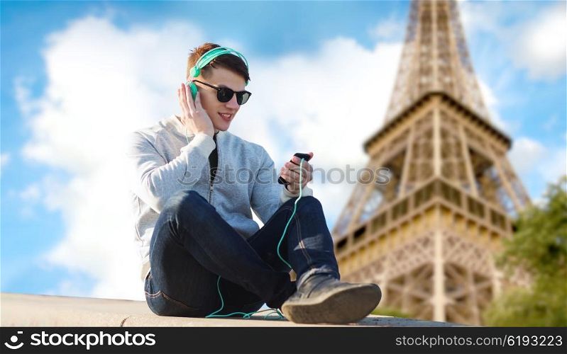 technology, travel, tourism and people concept - smiling young man or teenage boy in headphones with smartphone listening to music over paris eiffel tower background