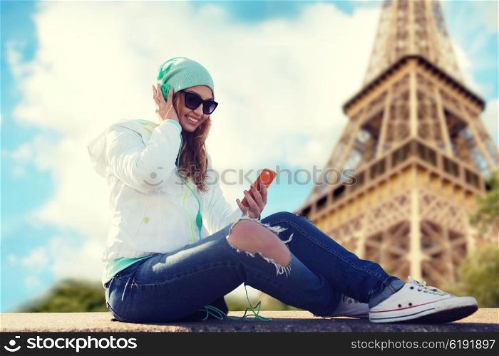 technology, travel, tourism and people concept - smiling young woman or teenage girl with smartphone and headphones listening to music over eiffel tower background