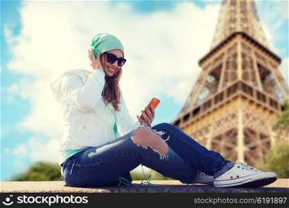 technology, travel, tourism and people concept - smiling young woman or teenage girl with smartphone and headphones listening to music over eiffel tower background