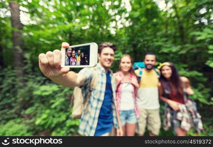 technology, travel, tourism, hike and people concept - close up of happy friends walking with backpacks taking selfie by smartphone in woods