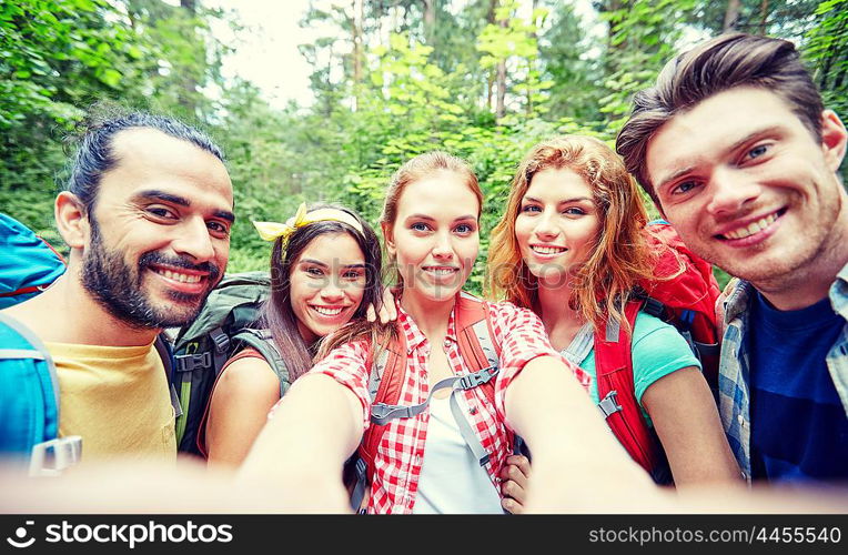 technology, travel, tourism, hike and people concept - group of smiling friends walking with backpacks taking selfie by smartphone or camera in woods