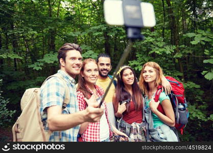 technology, travel, tourism, hike and people concept - group of smiling friends walking with backpacks taking picture by smartphone on selfie stick in woods