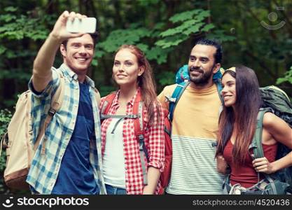 technology, travel, tourism, hike and people concept - group of smiling friends walking with backpacks taking selfie by smartphone in woods