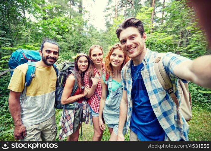 technology, travel, tourism, hike and people concept - group of smiling friends walking with backpacks taking selfie by smartphone or camera in woods