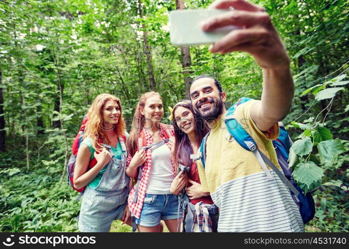 technology, travel, tourism, hike and people concept - group of smiling friends walking with backpacks taking selfie by smartphone in woods