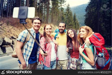 technology, travel, tourism, hike and people concept - group of smiling friends walking with backpacks taking picture by smartphone on selfie stick over woods and road background
