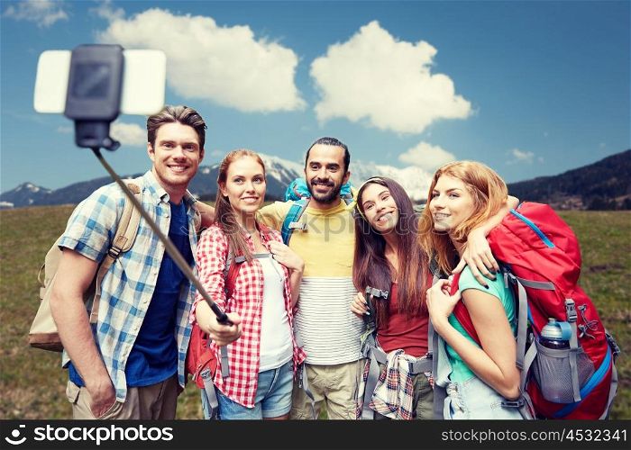 technology, travel, tourism, hike and people concept - group of smiling friends walking with backpacks taking picture by smartphone on selfie stick over natural background