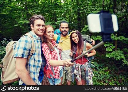 technology, travel, tourism, hike and people concept - group of smiling friends walking with backpacks taking picture by smartphone on selfie stick in woods