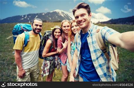 technology, travel, tourism, hike and people concept - group of smiling friends walking with backpacks taking selfie by smartphone or camera in over natural background