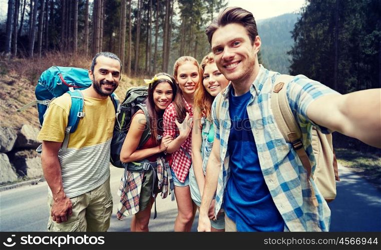 technology, travel, tourism, hike and people concept - group of smiling friends walking with backpacks taking selfie by smartphone or camera in over woods and road background
