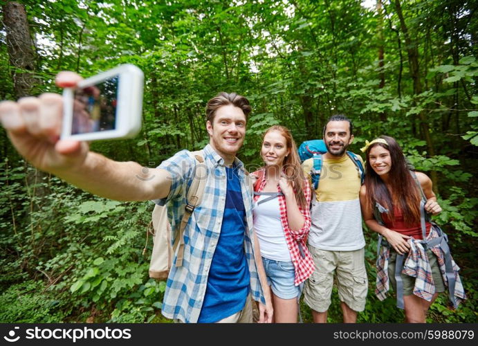 technology, travel, tourism, hike and people concept - group of smiling friends walking with backpacks taking selfie by smartphone in woods