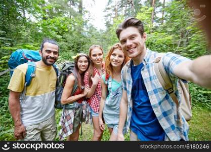 technology, travel, tourism, hike and people concept - group of smiling friends walking with backpacks taking selfie by smartphone or camera in woods
