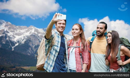 technology, travel, tourism, hike and people concept - group of smiling friends with backpacks taking selfie by smartphone over alps mountains background. friends with backpack taking selfie by smartphone. friends with backpack taking selfie by smartphone