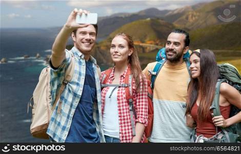 technology, travel, tourism, hike and people concept - group of smiling friends with backpacks taking selfie by smartphone over bixby creek bridge on big sur coast of california background. friends with backpack taking selfie by smartphone