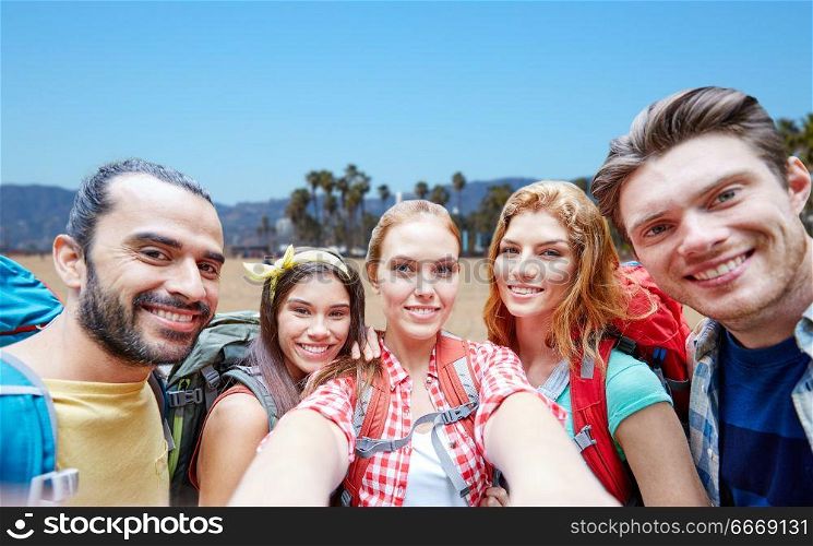 technology, travel, tourism, hike and people concept - group of smiling friends with backpacks taking selfie over venice beach background in california. friends with backpack taking selfie over beach. friends with backpack taking selfie over beach