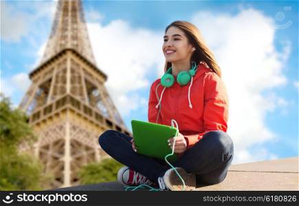 technology, travel, tourism, music and people concept - smiling young woman or teenage girl with tablet pc computer and headphones over paris eiffel tower background