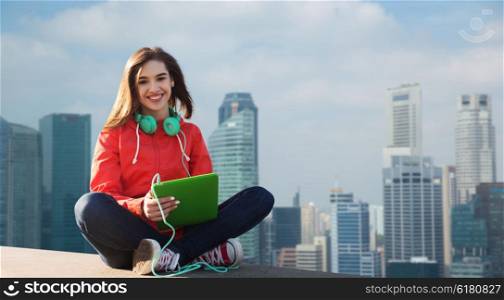 technology, travel, tourism, music and people concept - smiling young woman or teenage girl with tablet pc computer and headphones over singapore city skyscrapers background