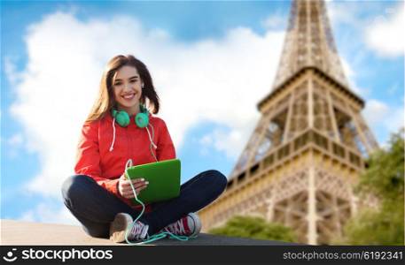 technology, travel, tourism, music and people concept - smiling young woman or teenage girl with tablet pc computer and headphones over paris eiffel tower background