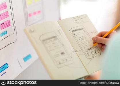 technology, user interface design and people concept - hand of ui designer or developer drawing smartphone sketches in notebook at office. ui designer with user interface sketch in notebook