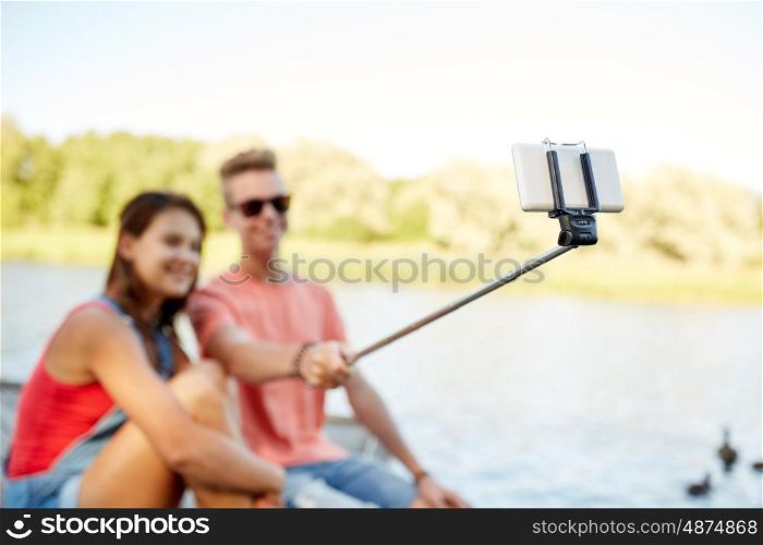 technology, vacation and people concept - happy teenage couple taking picture by smartphone selfie stick and sitting on river berth at summer