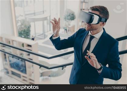 Technology VR. Businessman in virtual reality goggles working in office, dressed in suit standing on stairs and moving hands in air, touching objects in digital world, using innovations for business. Young businessman wearing virtual reality goggles working in office