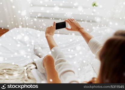 technology, winter, augmented rality and people concept - young woman in bed holding smartphone with black empty screen photographing at home bedroom over snow. woman photographing with smartphone in bet at home