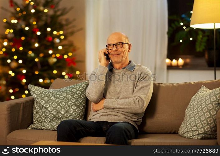 technology, winter holidays and communication concept - happy smiling senior man calling on smartphone at home in evening over christmas tree lights on background. senior man calling on phone at home on christmas