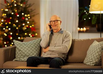 technology, winter holidays and communication concept - happy smiling senior man calling on smartphone at home in evening over christmas tree lights on background. senior man calling on phone at home on christmas