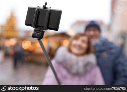 technology, winter holidays and gadgets concept - close up of smartphone on selfie stick taking picture of senior couple at christmas market on town hall square in tallinn, estonia. senior couple taking selfie at christmas market