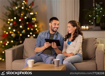 technology, winter holidays and people concept - happy couple using tablet computer at home in evening over christmas tree lights on background. happy couple using tablet pc at home on christmas