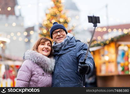 technology, winter holidays and people concept - happy senior couple taking picture by smartphone on selfie stick at christmas market on town hall square in tallinn, estonia. senior couple taking selfie at christmas market