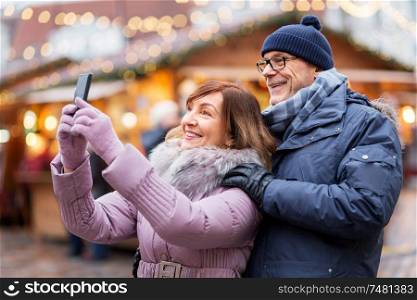 technology, winter holidays and people concept - happy senior couple taking selfie by smartphone at christmas market on town hall square in tallinn, estonia. senior couple taking selfie at christmas market