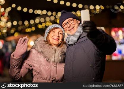 technology, winter holidays and people concept - happy senior couple taking selfie by smartphone at christmas market on town hall square in tallinn, estonia. senior couple taking selfie at christmas market