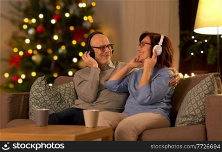 technology, winter holidays and people concept - happy senior couple with headphones listening to music at home in evening over christmas tree lights on background. senior couple with headphones listening to music
