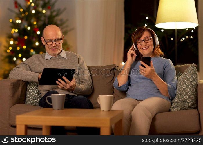 technology, winter holidays and people concept - happy senior couple with tablet pc computer, headphones and smartphone at home in evening over christmas tree lights on background. senior couple with gadgets at home on christmas