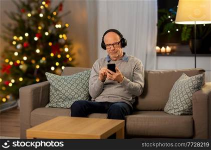 technology, winter holidays and people concept - happy senior man with smartphone and headphones listening to music at home in evening over christmas tree lights background. senior man with smartphone and headphones at home