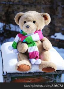 teddy bear in a scarf sits on a stump in the middle of white snow on a winter day