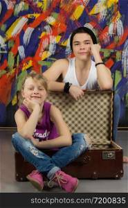 Teen boy wearing tank top and fashion cap and girl sitting in empty suitcase and smiling at camera