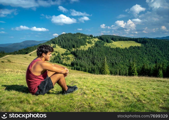 Teen Boy with Pleasure Sitting on the Fresh Green Grass on the Hill of Carpathian Mountains. Enjoying Beautiful Landscape. Summer Activity. Trekking. Ukraine.