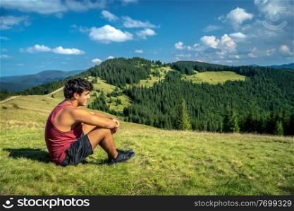Teen Boy with Pleasure Sitting on the Fresh Green Grass on the Hill of Carpathian Mountains. Enjoying Beautiful Landscape. Summer Activity. Trekking. Ukraine.