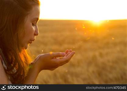 teen girl blowing at her palms filled with wheat grains standing at wheat field at sunset time