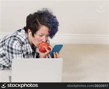 Teen girl eating apple while looking at cell phone with computer in forefront while lying down listening to music at home.